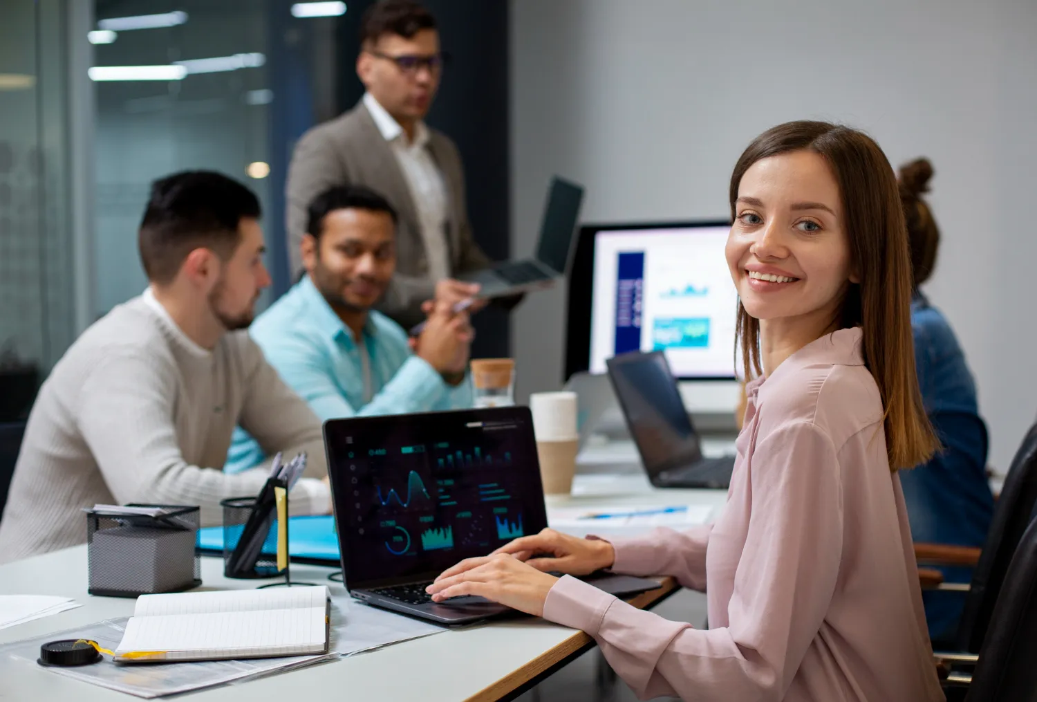 Smiling woman at a team meeting, working on data analysis using a laptop, with colleagues discussing in the background.