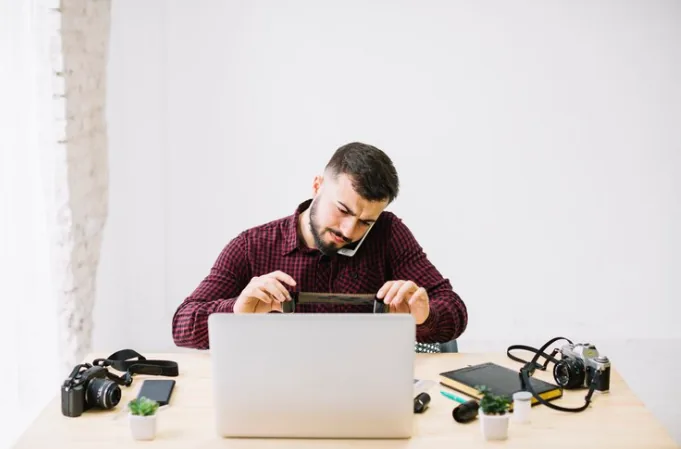 Man sits at a desk using a laptop, surrounded by cameras, lenses, notebooks, and small potted plants.