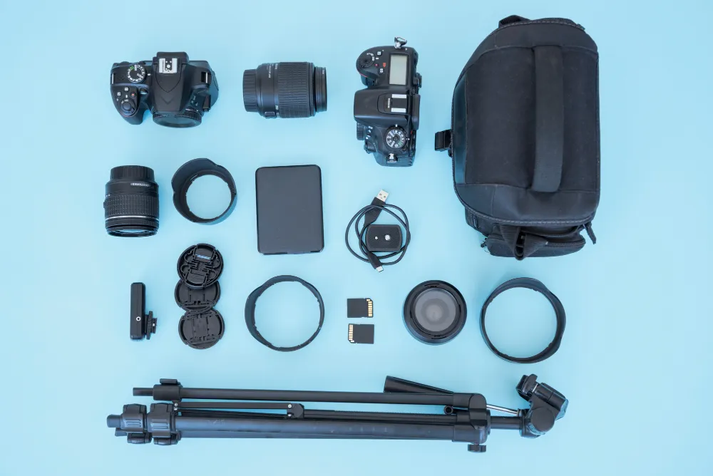 Camera equipment, lenses, tablet, cables, memory cards, filters, backpack, and tripod arranged on a blue background.