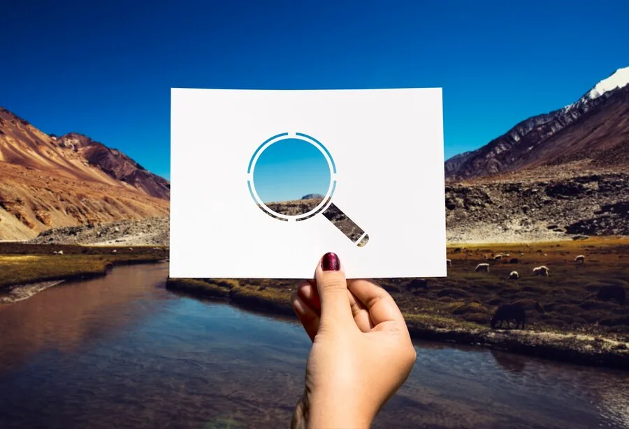 Hand holds a white card with a magnifying glass graphic against a mountainous landscape and winding river.
