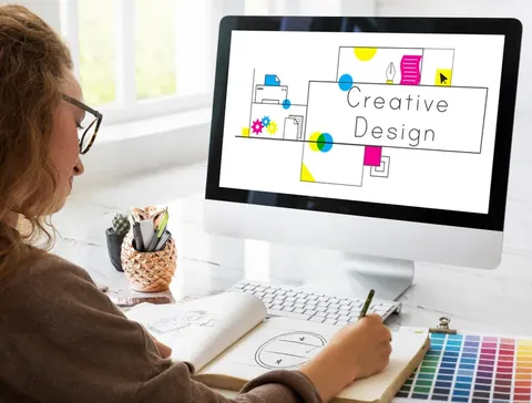 Designer sketches on paper beside a computer displaying “Creative Design,” with colored pencils, stationery, and color swatches nearby.
