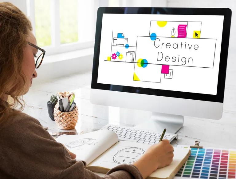 Designer sketches on paper beside a computer displaying “Creative Design,” with colored pencils, stationery, and color swatches nearby.